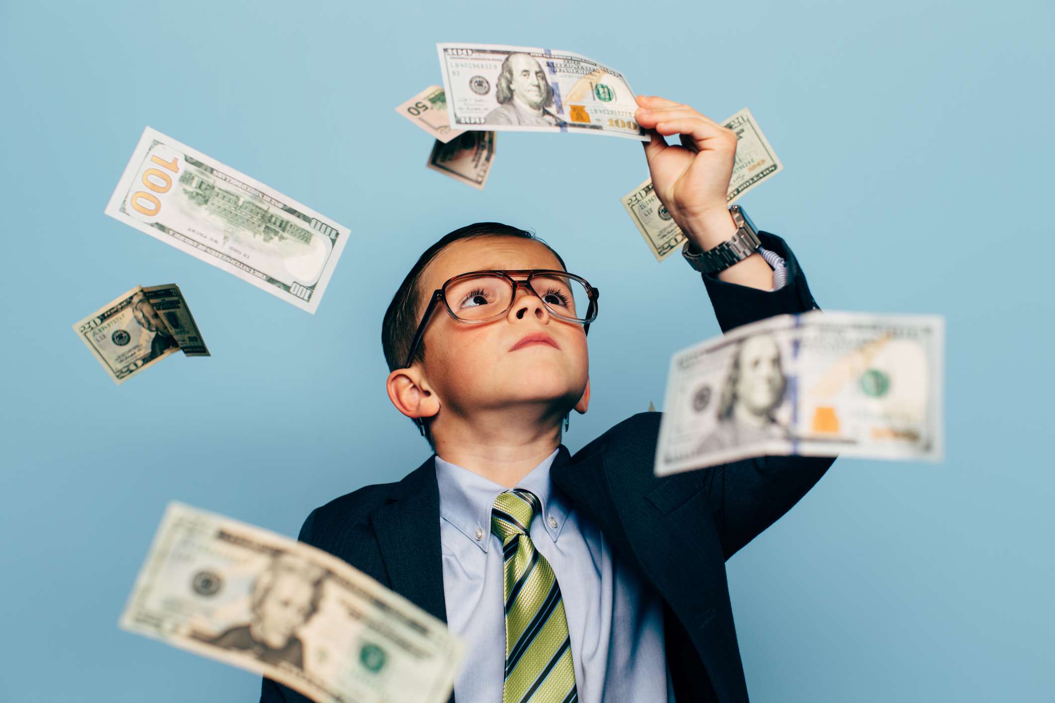 A young boy accountant wearing glasses and suit watches U.S. currency while more falls from above. He is looking closely at Ben Franklin.