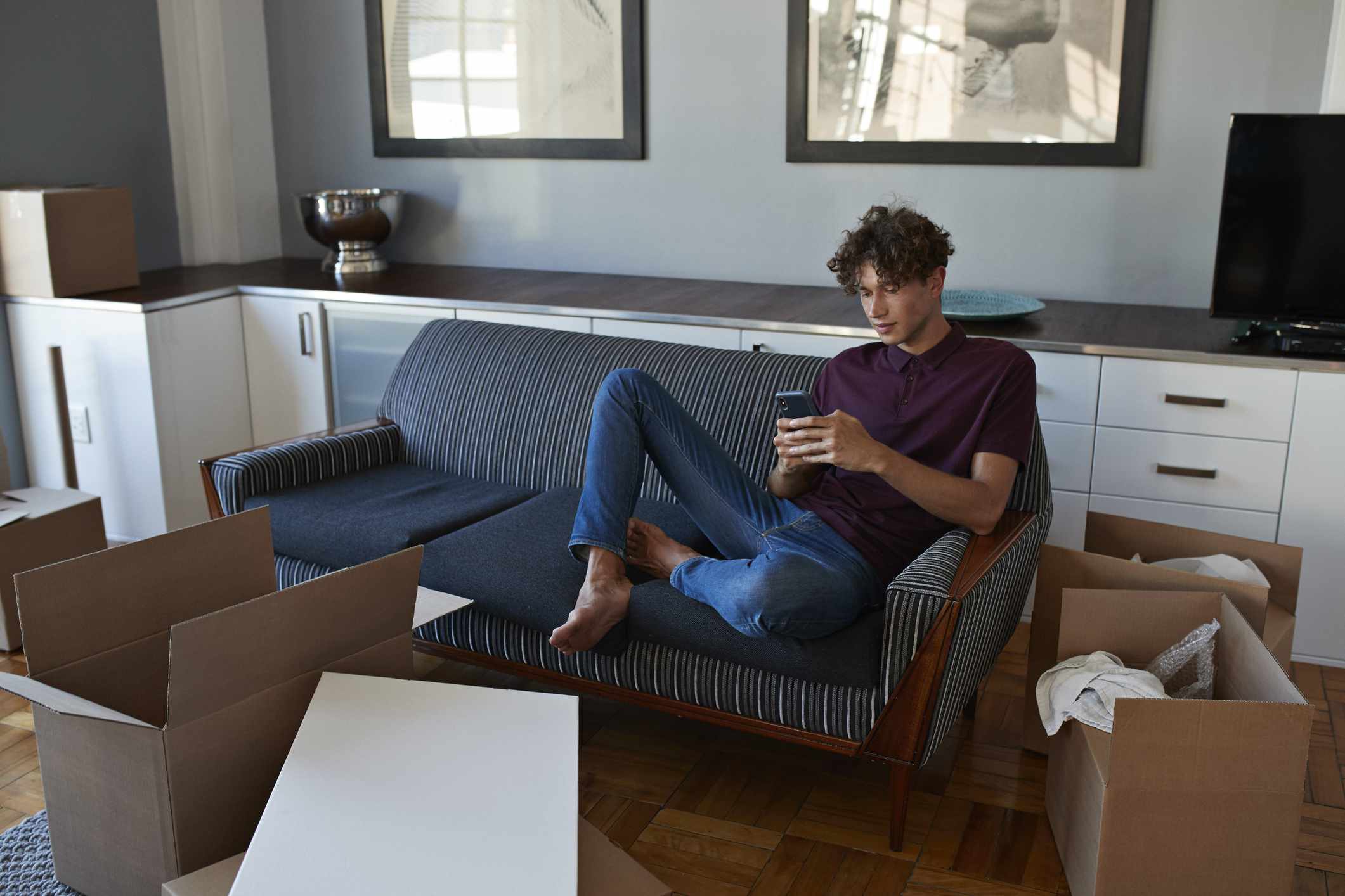 A renter sits on a couch texting, surrounded by open cardboard boxes in his new apartment