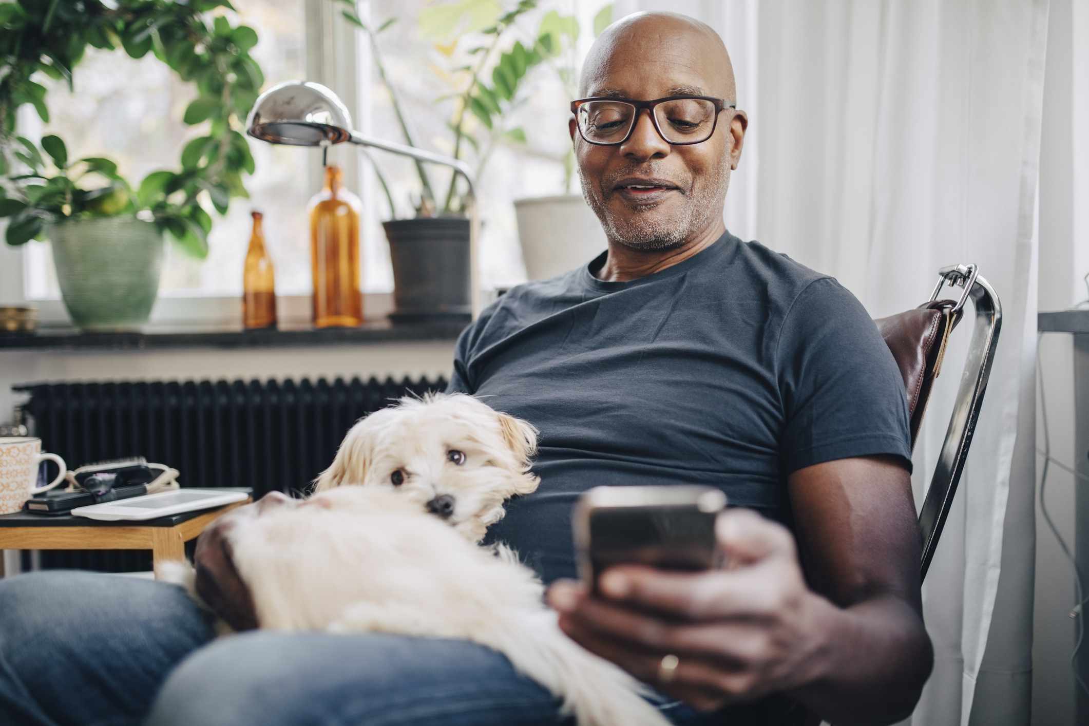Smiling retired senior male using smartphone while sitting with dog in room at home