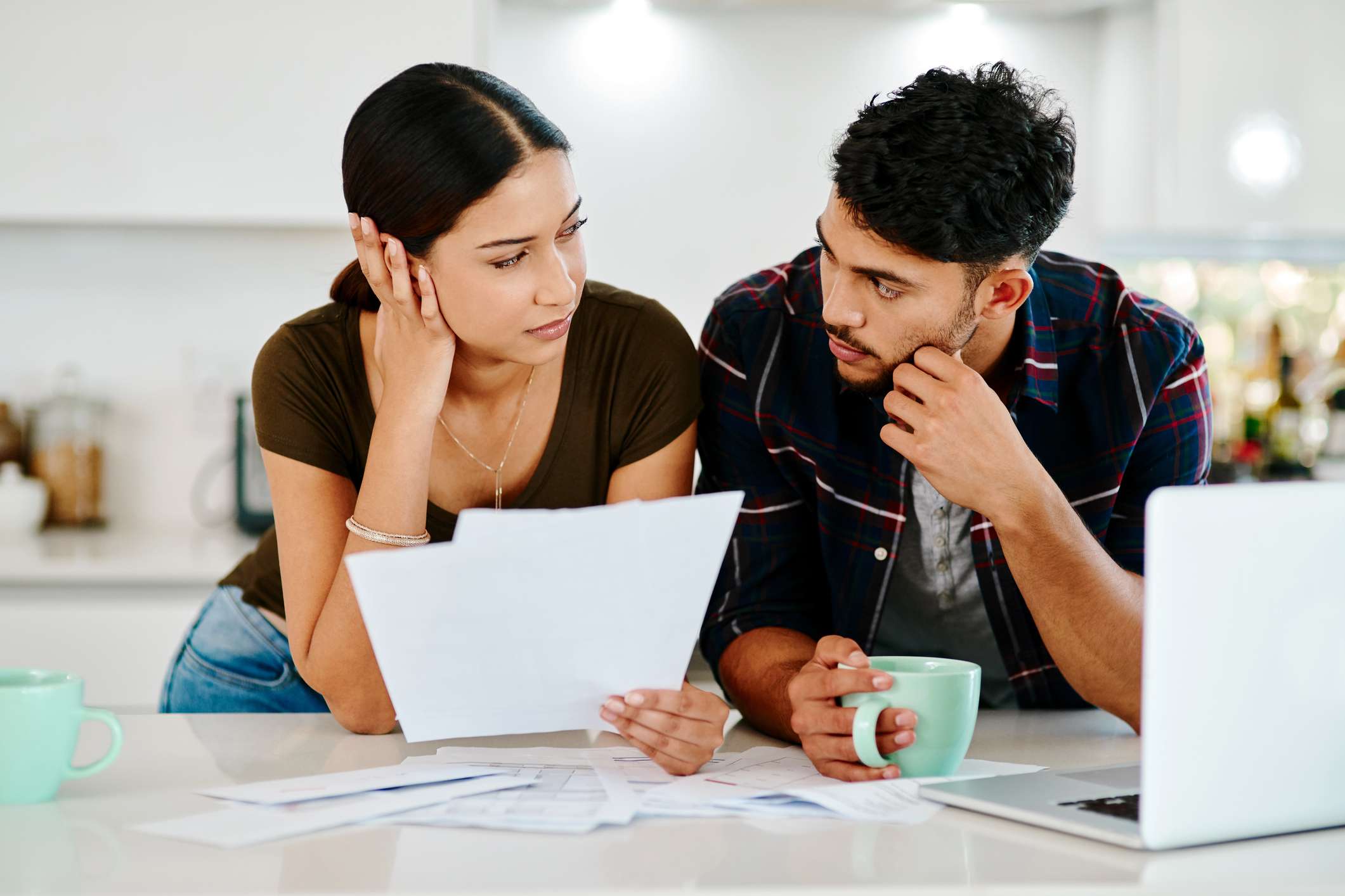 A stressed young couple look at one another, holding papers while drinking coffee in their kitchen, while deciding whether to file a home insurance claim.