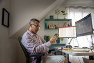 Man sitting down at computer desk looking at charts on his phone and two monitors.