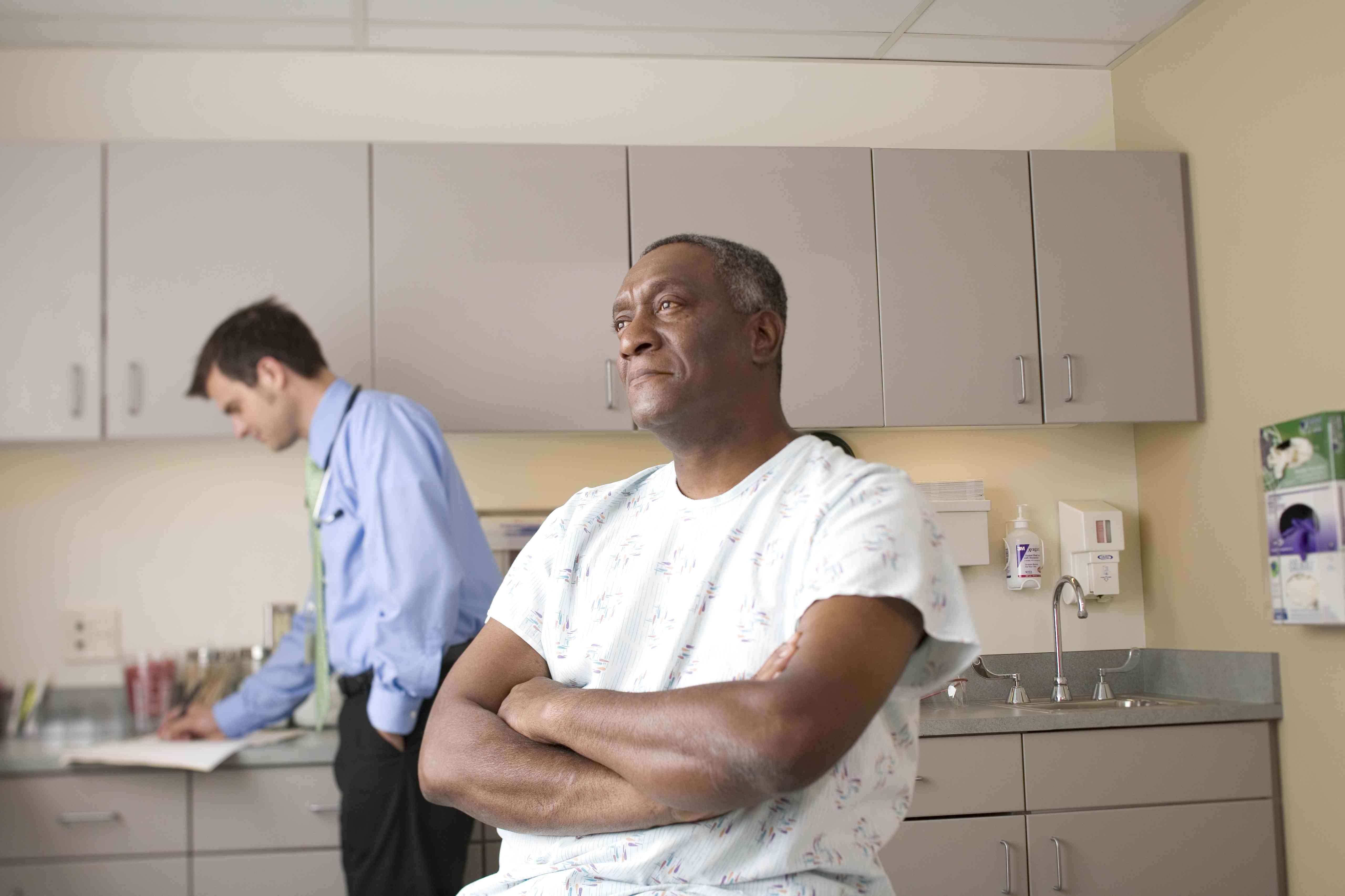 Black man with arms crossed in patient gown while White doctor writes in file