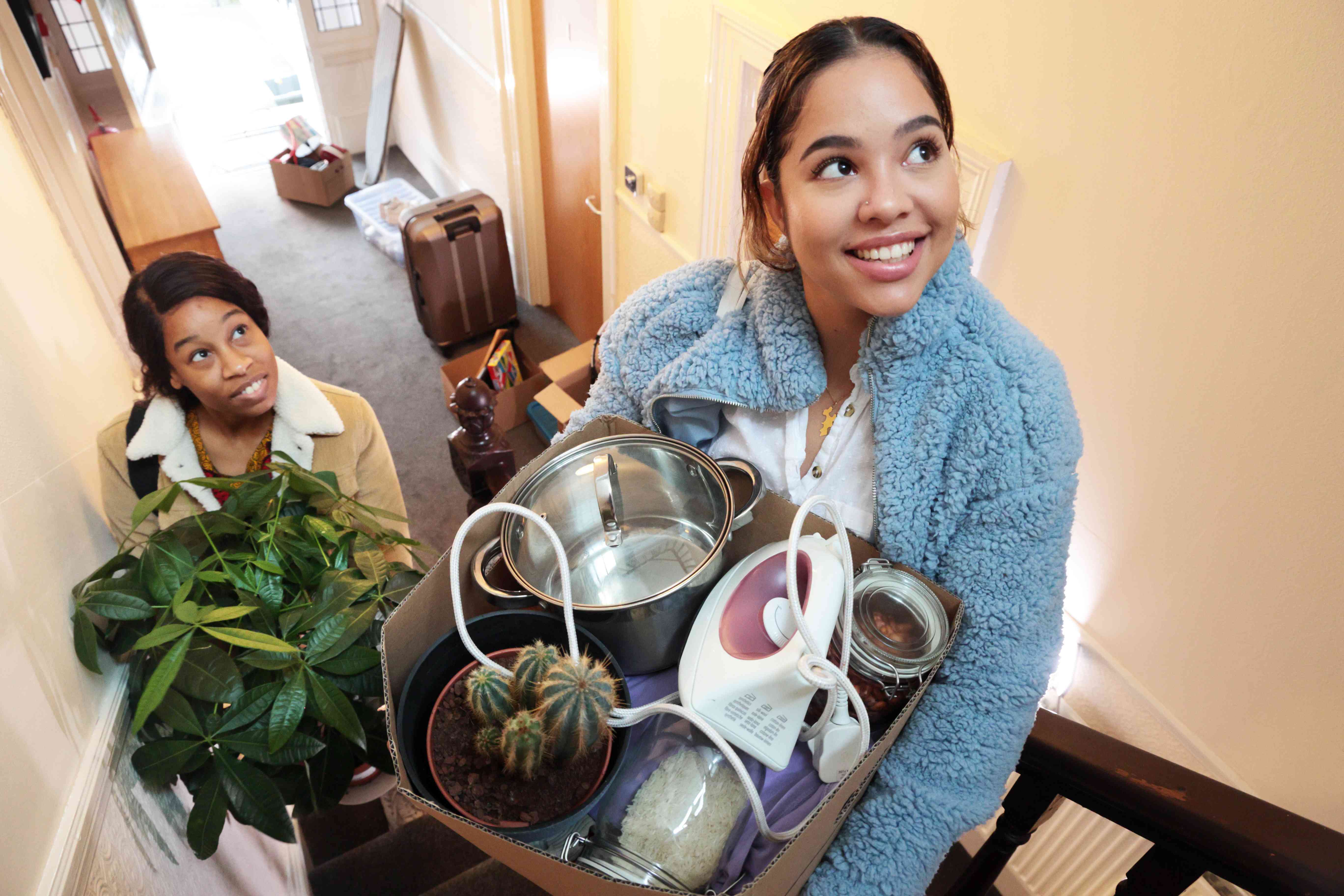 Two young women carry plants and a box of belongings up some interior stairs